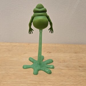 May include: A green, stylized figurine of a creature with a bulbous body, small arms, and a head with stacked segments. The figure is mounted on a slender stem attached to a star-shaped base. The figurine is on a wooden surface.