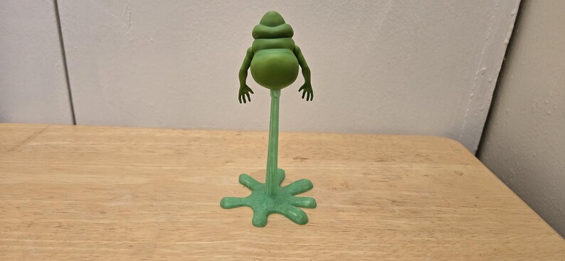 May include: A green, stylized figurine of a creature with a bulbous body, small arms, and a head with stacked segments. The figure is mounted on a slender stem attached to a star-shaped base. The figurine is on a wooden surface.