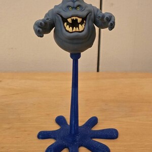 May include: A blue, cartoon-like figure with a wide grin and outstretched arms, mounted on a blue stand. The figure has a bulbous body and a textured head. The stand has a star-shaped base.