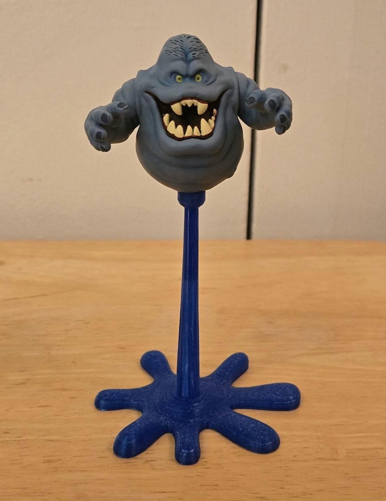 May include: A blue, cartoon-like figure with a wide grin and outstretched arms, mounted on a blue stand. The figure has a bulbous body and a textured head. The stand has a star-shaped base.