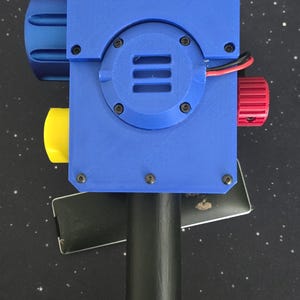 May include: A handheld device with a blue rectangular housing, a black cylindrical handle, and a circular blue speaker grille. It features a large blue knob on the left, a yellow knob on the bottom left, and a red knob on the right with visible red and black wires.