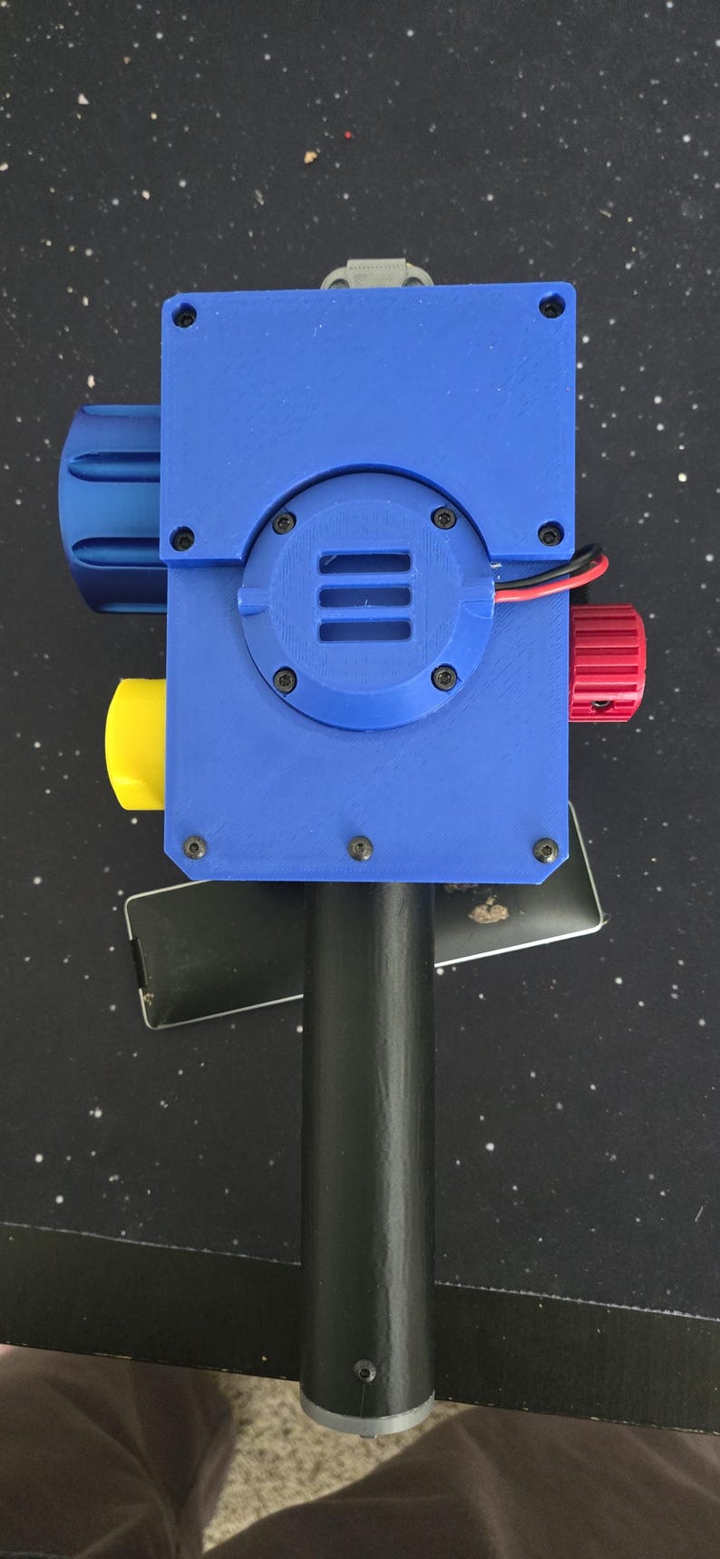 May include: A handheld device with a blue rectangular housing, a black cylindrical handle, and a circular blue speaker grille. It features a large blue knob on the left, a yellow knob on the bottom left, and a red knob on the right with visible red and black wires.