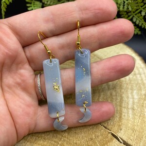 May include: A pair of handmade earrings featuring a rectangular, blue and white marbled design with gold flakes. Each earring has a gold-colored hook and a small, gray crescent moon charm attached to the bottom.