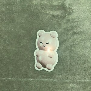 May include: A pink gummy bear sticker with a cartoon face. The bear has a small smile and closed eyes.