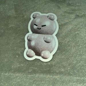 May include: A sticker of a light pink gummy bear with a white outline. The gummy bear has a small smile and a few white dots on its body.