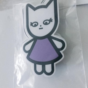 Op de afbeelding: Een witte sticker met een zwarte omtrek van een kat die een paarse jurk draagt. De kat heeft een eenvoudig gezicht met zwarte ogen en een kleine mond.