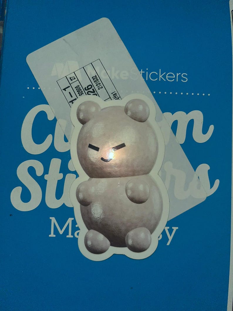 May include: A white gummy bear sticker with a black face and a slight smile. The sticker is on a blue background with the text "Custom Stickers" and "Make Stickers" in white.