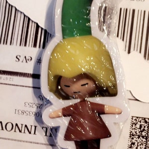 May include: A white sticker with a cartoon illustration of a person with yellow hair wearing a green hat and a brown dress. The sticker is wrapped in plastic.