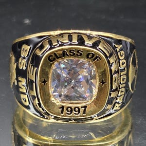 Könnte beinhalten: Ein goldfarbener Ring mit einem zentralen, kissenförmigen, klaren Stein. Die schwarze Emaille-Vorderseite trägt die Inschrift "CLASS OF 1997" und "UNIVERSITY". Die Seiten zeigen den Text "BS MD" und "RADIOLOGY" in goldener Schrift auf schwarzem Grund.