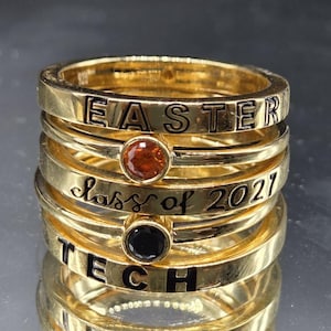 Könnte beinhalten: Ein Stapel goldfarbener Ringe mit eingraviertem Text und eingesetzten Steinen. Der oberste Ring trägt das Wort EASTER, gefolgt von einem Ring mit einem orangefarbenen Stein, einem Ring mit dem Text class of 2021, einem Ring mit schwarzem Stein und TECH.