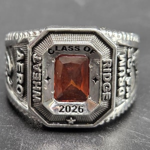 Könnte beinhalten: Ein silberfarbener Klassenring mit einem rechteckigen, facettierten bernsteinfarbenen Stein in der Mitte. Der Ring ist mit dem Text "CLASS OF 2026", "WHEAT RIDGE", "AERO" und "DRUM MAJOR" um die zentrale Steinfassung herum graviert.