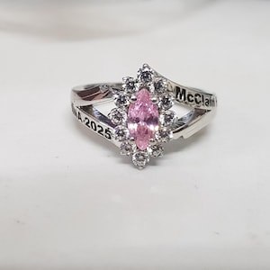 Może przedstawiać: Srebrny pierścionek z różowym kamieniem centralnym w kształcie markizy, otoczonym mniejszymi, przezroczystymi kamieniami. Na obrączce widnieje napis "McClain 2025".