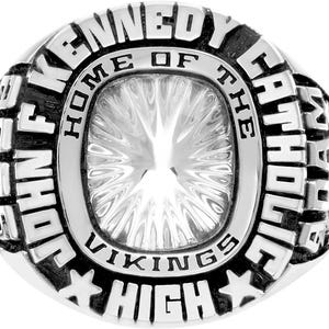 May include: A silver class ring with the text "JOHN F. KENNEDY CATHOLIC HIGH" and "HOME OF THE VIKINGS". The ring has a central, clear, faceted stone and star accents.