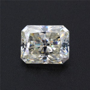 Pode incluir: Um diamante de corte radiante em forma retangular. A gema tem múltiplas facetas que refletem a luz, criando um efeito brilhante. O diamante está sobre um fundo escuro.