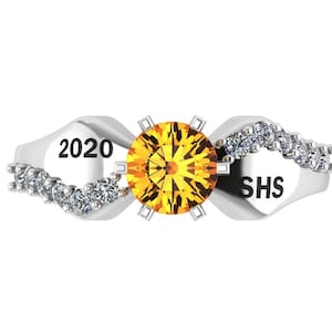 May include: A silver ring with a large, round, yellow gemstone at the center. The ring has a wave-like design with small, clear stones and the text "2020" and "SHS" on either side of the center stone.