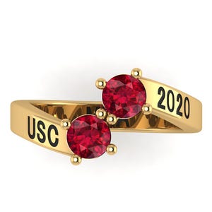 May include: A gold-colored ring with two red gemstones. The ring has the letters "USC" and the number "2020" engraved on the band. The gemstones are round and set in a gold bezel.