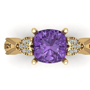 Könnte beinhalten: Ein goldener Ring mit einem großen, quadratischen, lila Edelstein in der Mitte. Der Ring ist mit kleinen Diamanten verziert und trägt die Inschriften "2021" und "MS D" an den Seiten. Der Ring ist vor einem weißen Hintergrund platziert.