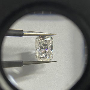 Op de afbeelding: Een radiant-geslepen diamant, vastgehouden door een pincet, bekeken door een vergrootglas. De diamant is rechthoekig met een briljant, facetrijk oppervlak dat licht reflecteert. De steen lijkt kleurloos, met een hoge mate van helderheid, tegen een witte achtergrond.