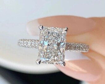 May include: A radiant-cut diamond engagement ring. The ring features a large, rectangular-shaped diamond as the centerpiece, with smaller diamonds set along the band. The ring is set in a white metal, likely platinum or white gold.