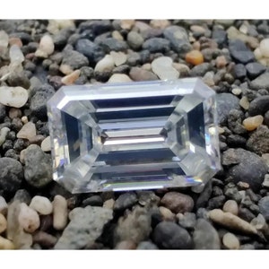 Pode incluir: Uma gema clara, com corte esmeralda, de forma retangular e bordas facetadas. A gema está sobre um fundo de pequenas pedras cinzentas e bege. As facetas refletem a luz, criando um efeito brilhante.