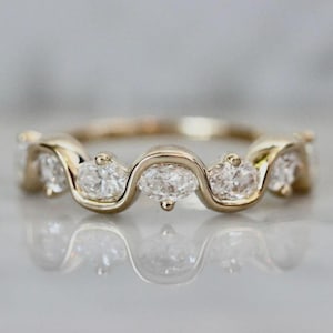 Puede incluir: Un anillo de banda de oro con una serie de piedras preciosas claras de corte ovalado. Las piedras preciosas están engastadas en un patrón ondulado, creando un diseño único y elegante. El anillo se muestra sobre una superficie reflectante.