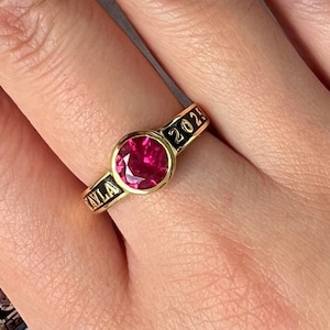 Könnte beinhalten: Ein goldener Ring mit einem großen, runden, roten Edelstein. Auf dem Ringband sind der Name "KYLA" und das Jahr "2023" in Schwarz eingraviert. Der Hintergrund ist unscharf.