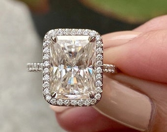 May include: A radiant-cut diamond ring with a halo of smaller diamonds. The rectangular center stone is surrounded by a square halo of pave-set diamonds. The ring's band also features pave-set diamonds, adding to its sparkle.