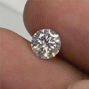 May include: A round, brilliant-cut diamond with multiple facets, reflecting light in a spectrum of colors. The gemstone is held between fingers, showcasing its clarity and sparkle. The diamond's cut maximizes its brilliance.