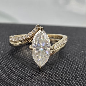 May include: A gold ring featuring a large marquise-cut center stone held by four prongs. The band has a bypass design with two sweeping arms, each set with a row of small round accent stones. The inner band is clearly stamped with the text 10K.