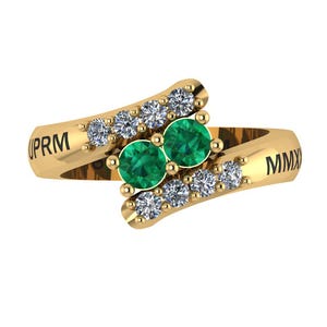 Może przedstawiać: Złoty pierścionek o konstrukcji typu bypass. Pierścionek posiada dwa szmaragdowe zielone kamienie szlachetne i kilka małych, przezroczystych kamieni szlachetnych. Na obrączce pierścionka wygrawerowano tekst "PRM" i "MMX".