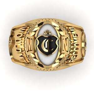 Puede incluir: Un anillo dorado con un diseño central de escudo negro y blanco. El anillo presenta las palabras "WORLD" y "RECOMMEND" en los laterales, con "M" y "N" en la parte delantera. Una corona se encuentra encima del escudo.