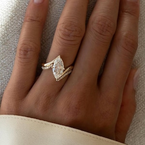 Könnte beinhalten: Ein goldener Verlobungsring mit einem Diamanten im Marquise-Schliff als Mittelpunkt. Das Band hat ein zartes, geschwungenes Design mit kleineren Akzentsteinen. Der Ring wird an einer Hand gezeigt und unterstreicht seinen eleganten und modernen Stil.