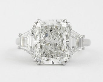 May include: A three-stone ring featuring a large rectangular radiant-cut center diamond held by eight prongs, flanked by two smaller trapezoid-cut side diamonds, all set on a polished silver-toned metal band against a plain white background.