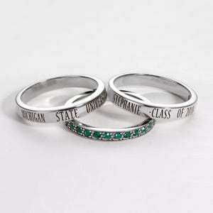 Könnte beinhalten: Drei silberne Ringe. Zwei Ringe tragen eingravierte Texte: "MICHIGAN STATE UNIVERSITY" und "STEPHANIE - CLASS OF 2018". Der dritte Ring ist mit kleinen grünen Edelsteinen besetzt. Alle Ringe auf weißem Hintergrund.
