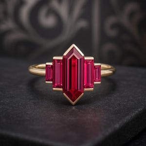Könnte beinhalten: Ein goldener Ring mit einem länglichen, rubinroten Edelstein in der Mitte, flankiert von kleineren, rechteckigen Rubinen. Der Ring hat ein geometrisches Design mit einem goldenen Band und einer Fassung im Art-Deco-Stil.