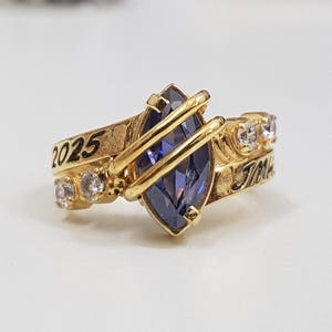 Puede incluir: Un anillo de clase dorado con una piedra preciosa azul en forma de marquesa, rodeada de pequeñas piedras blancas. El anillo tiene el año "2025" grabado en un lado y la palabra "Jim" grabada en el otro.