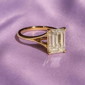 Könnte beinhalten: Ein goldener Ring mit einem großen, rechteckig geschliffenen Diamanten. Das Band des Rings teilt sich in zwei zarte Zinken, die den Diamanten halten. Der Ring ist vor einem weichen, lila Hintergrund platziert.