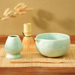 May include: A matcha tea set comprising a pale green ceramic bowl, a whisk, a whisk holder, and a bamboo scoop. The set is displayed on a bamboo mat, against a neutral backdrop. The bowl has a spout for pouring.