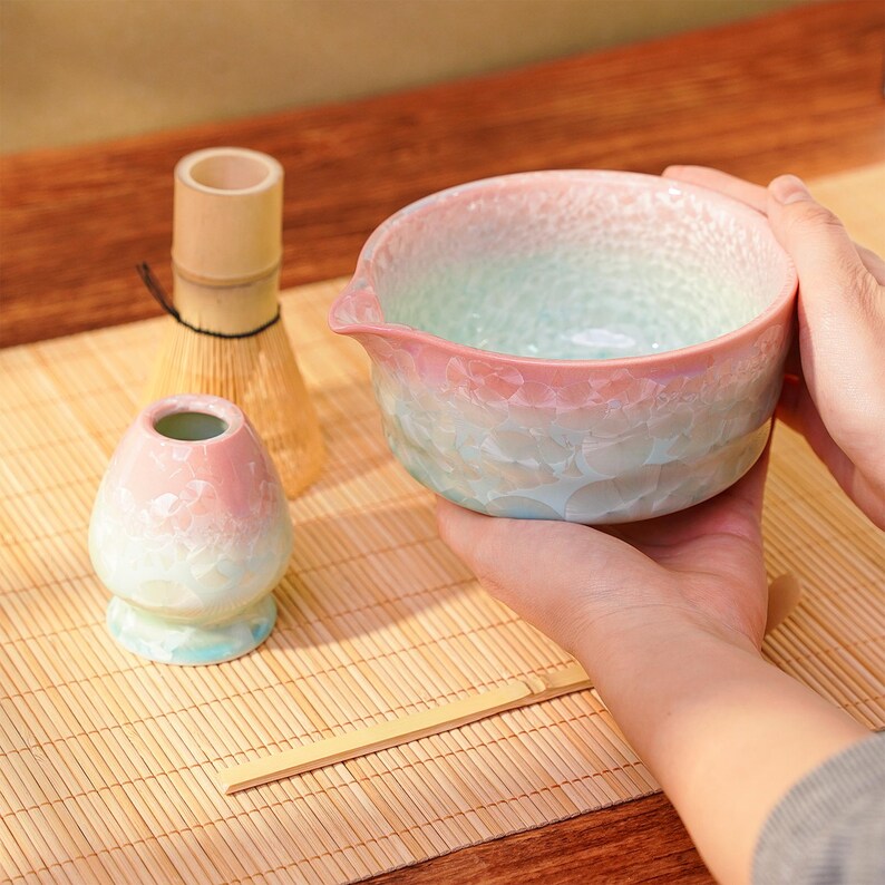 Pu&ograve; includere: Un set da t&egrave; matcha con una ciotola sfumata, una frusta, un piccolo contenitore e un cucchiaio di bamb&ugrave;. La ciotola &egrave; tenuta tra le mani e presenta una combinazione di colori rosa, verde e blu. La frusta e il cucchiaio sono in bamb&ugrave;.