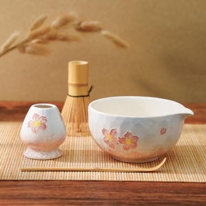 Op de afbeelding: Een matcha-theeset met een witte keramische kom met roze bloemmotieven, een bamboe klopper, een kleine keramische houder en een bamboelepel. De set is geplaatst op een bamboemat, met een neutrale achtergrond.