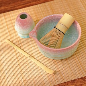 Pu&ograve; includere: Un set da t&egrave; matcha con una ciotola in ceramica rosa e blu, un frullino in bamb&ugrave;, un piccolo contenitore in ceramica e un cucchiaio in bamb&ugrave;. Il set &egrave; disposto su un tappetino di bamb&ugrave;, creando un'estetica serena e tradizionale.