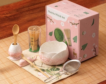 Può includere: Un set da 7 pezzi per matcha in una scatola rosa con illustrazioni floreali. Include una ciotola in ceramica bianca e rosa, un frustino in bambù in una custodia trasparente, un cucchiaio in bambù, un supporto, un poggia-frustino e un colino.