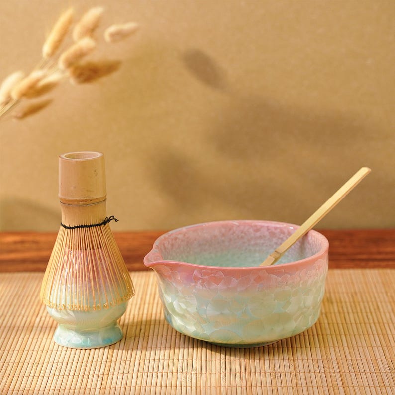 Pu&ograve; includere: Un set da t&egrave; matcha con un frullino di bamb&ugrave;, una ciotola di ceramica con beccuccio e un cucchiaio di bamb&ugrave;. La ciotola ha una sfumatura rosa e verde, e il frullino ha una sfumatura marrone chiaro e verde. Il set &egrave; su un tappetino di bamb&ugrave;.