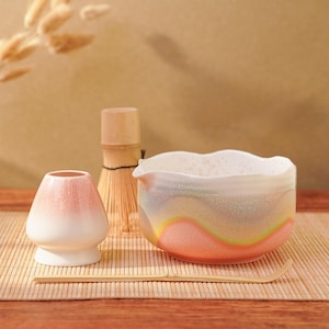 Può includere: Un set da tè matcha con una ciotola chawan in ceramica bianca e rosa con bordo ondulato, un frullino di bambù, una piccola tazza da tè in ceramica rosa e bianca e un cucchiaio di bambù. Il set è disposto su un tappetino di bambù.
