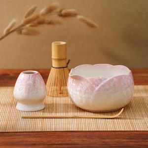 Puede incluir: Un juego de té matcha con un cuenco de cerámica rosa y blanco, un batidor, una pequeña taza de cerámica y una cuchara de bambú. El cuenco tiene un diseño floral. El juego está dispuesto sobre una esterilla de bambú.