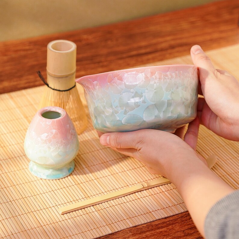 Pu&ograve; includere: Una ciotola da matcha in ceramica dai colori pastello, con un beccuccio, tenuta in mano. La ciotola &egrave; rosa, blu e bianca con una superficie strutturata. Sono visibili anche un portabattitore matcha e un frustino di bamb&ugrave; abbinati, il tutto su un tappetino di bamb&ugrave;.