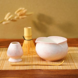 Może przedstawiać: Zestaw do herbaty matcha składający się z różowo-białej ceramicznej miski, bambusowej trzepaczki, małego ceramicznego uchwytu i bambusowej łyżki. Miska ma brzeg w kształcie płatka. Zestaw jest ułożony na bambusowej macie.
