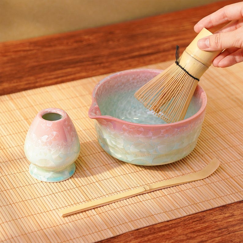 Pu&ograve; includere: Un set da t&egrave; matcha con una ciotola in ceramica rosa e blu, un frullino in bamb&ugrave;, un cucchiaio in bamb&ugrave; e un contenitore per t&egrave; in ceramica. La ciotola ha una superficie strutturata. Il frullino &egrave; sopra la ciotola.