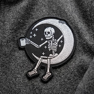 May include: Embroidered patch featuring a skeleton sitting on a crescent moon, holding a mug. The patch is black with white and gray details, including stars. The patch is attached to a gray fabric.