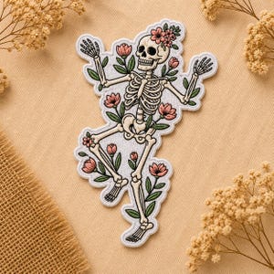 May include: Embroidered patch featuring a dancing skeleton adorned with pink flowers and green leaves. The skeleton has a floral crown and is surrounded by more flowers. The patch has a white border and is on a beige background.